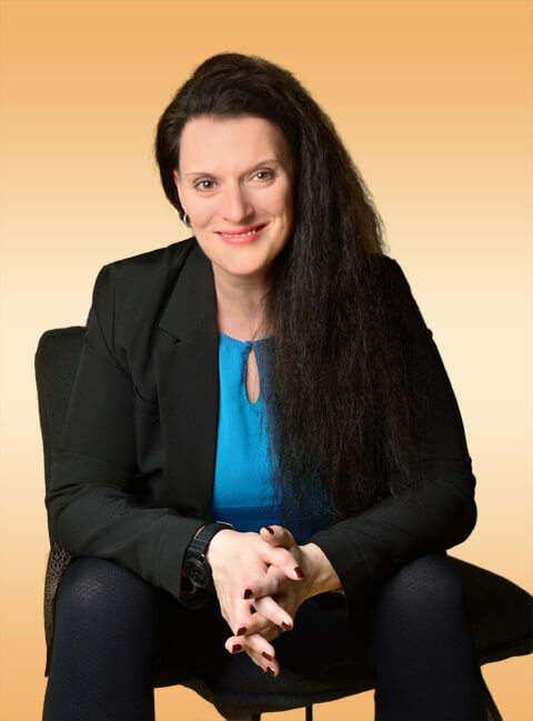 Cornelia Leclerc, Stress Coach, sitzt lächelnd, mit auf den Knien aufgestützten Ellenbogen und verschränkten Fingern auf einem Stuhl